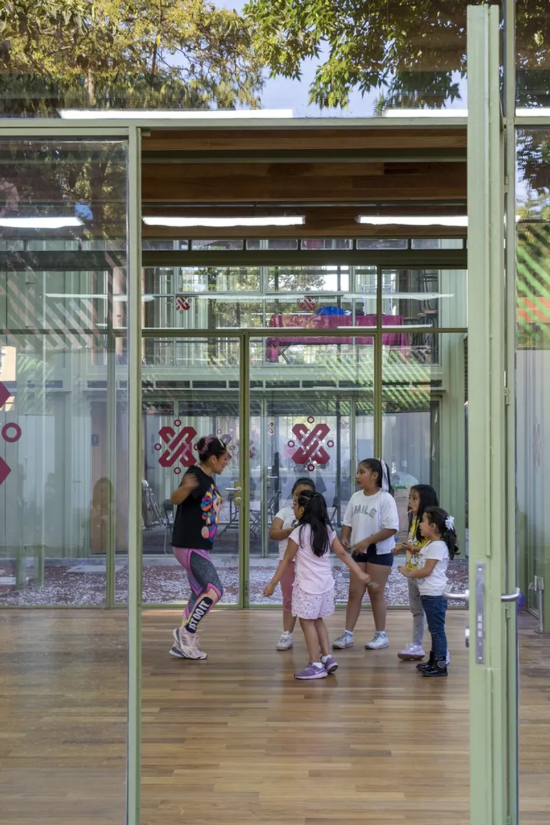 Open-air pavilion with green steel frame and timber floor where children gather in dappled sunlight