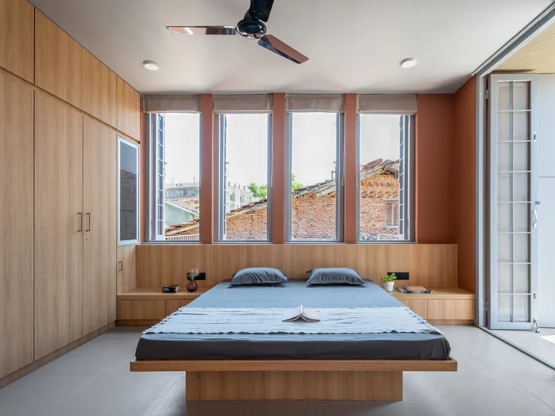 Bedroom with platform bed and timber window seat beneath four vertical windows overlooking terracotta rooftops