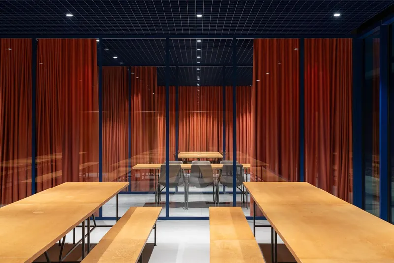 Interior dining hall with timber benches and glazed partitions with orange curtains under a black grid ceiling