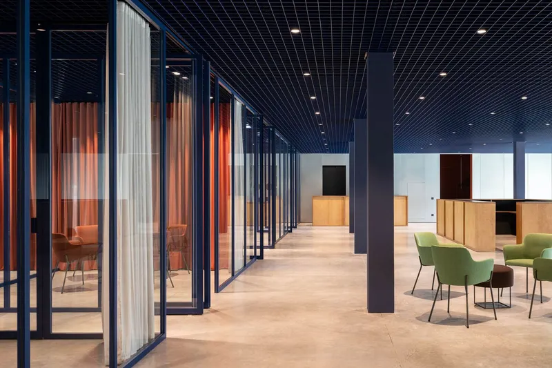 Corridor with black grid ceiling leading past glazed meeting rooms with curtains and green lounge chairs