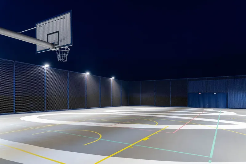 Outdoor basketball court with painted court lines and perimeter fencing illuminated at night