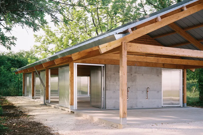 Covered pavilion with exposed timber rafters, concrete walls, and translucent sliding panels surrounded by trees