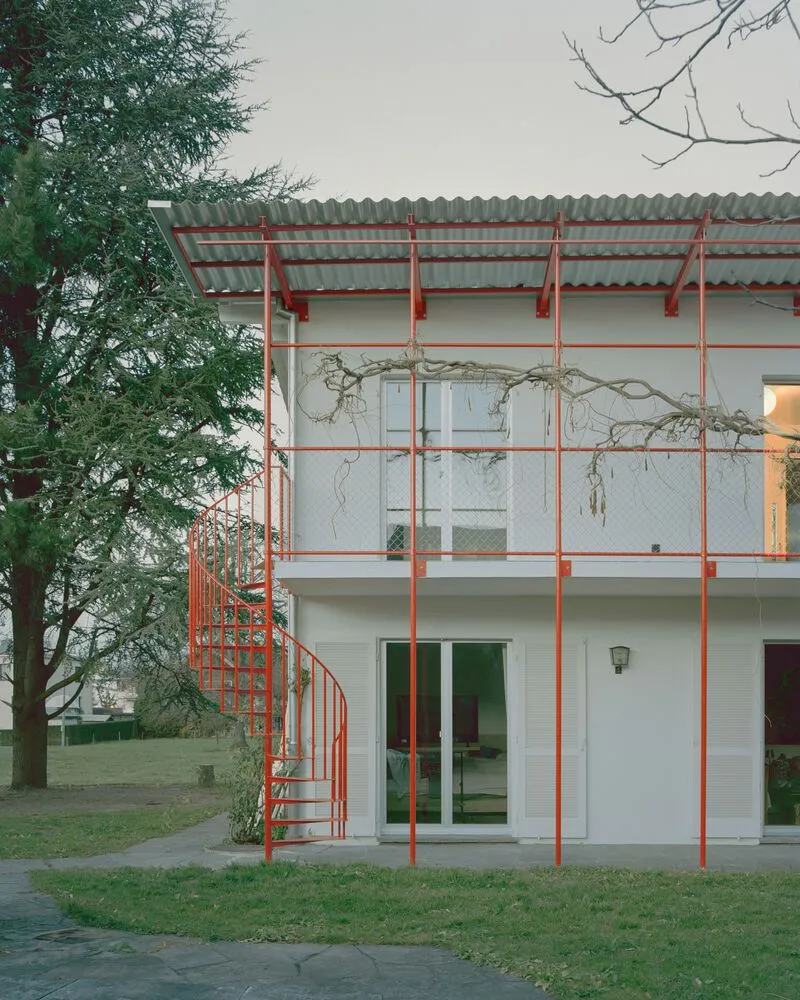 Red steel spiral staircase connecting two levels of the balcony structure under vine-draped branches at dusk