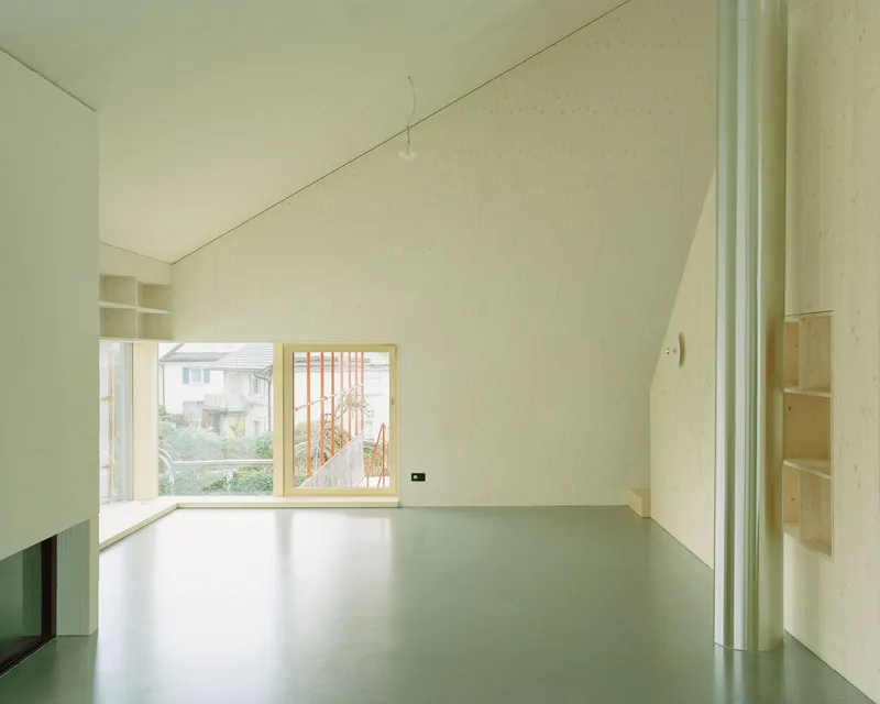 Interior room with sloped plywood ceiling, pale green resin floor and window overlooking the red exterior framework