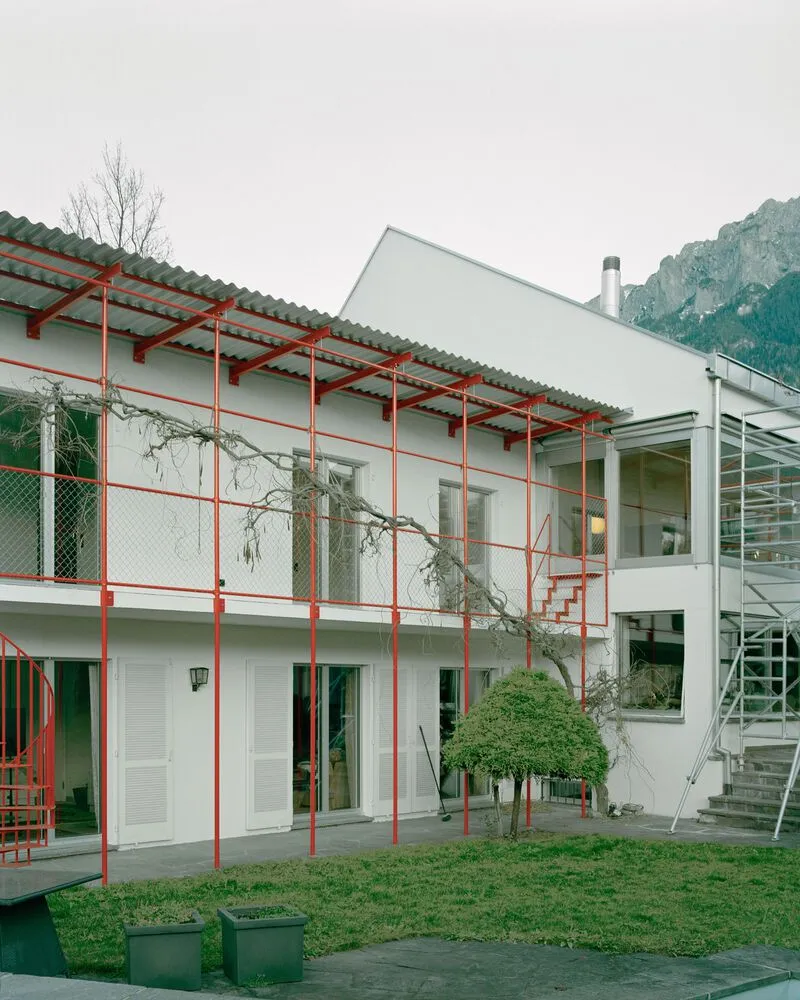 Courtyard view of the red steel balcony structure with corrugated canopy and bare trees against a mountain backdrop