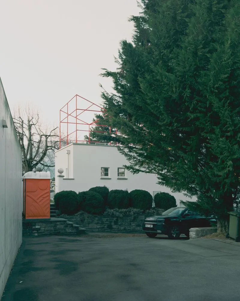 Street view showing the red skeletal rooftop structure above white walls with evergreen trees and parked car