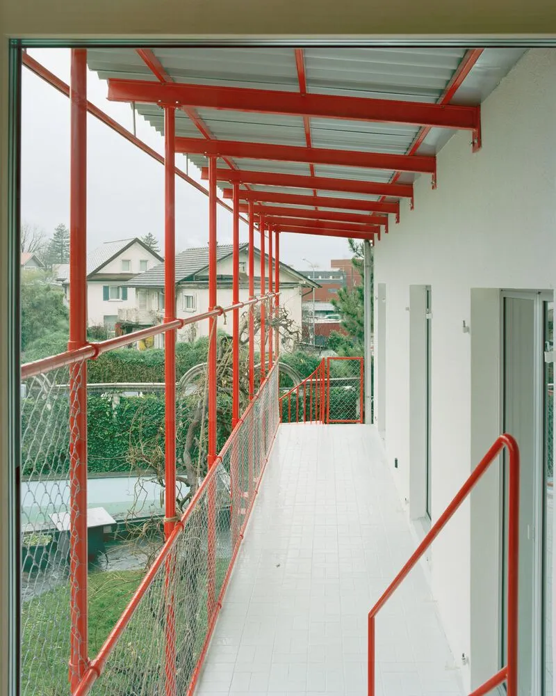Exterior walkway with red steel frame and mesh railing overlooking a residential neighborhood