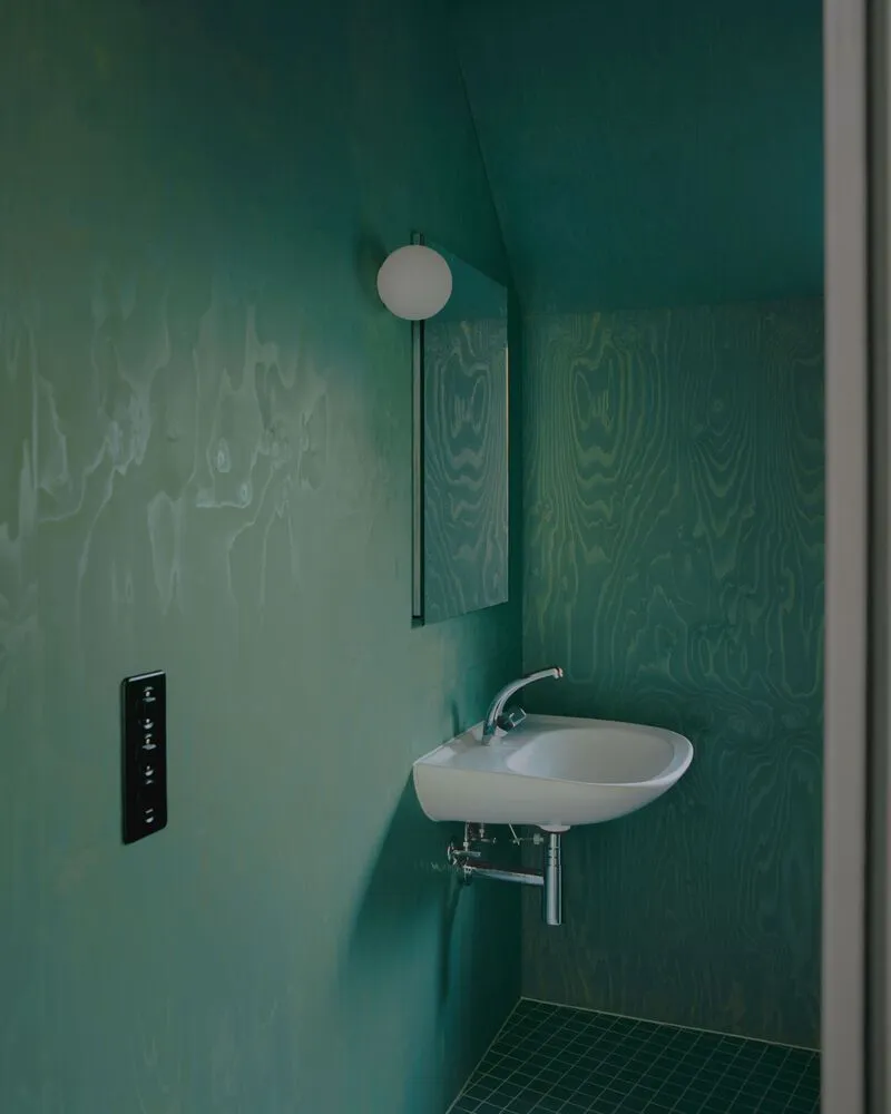 Corner of green plywood-paneled bathroom showing circular mirror and wall-mounted sink with tiled floor