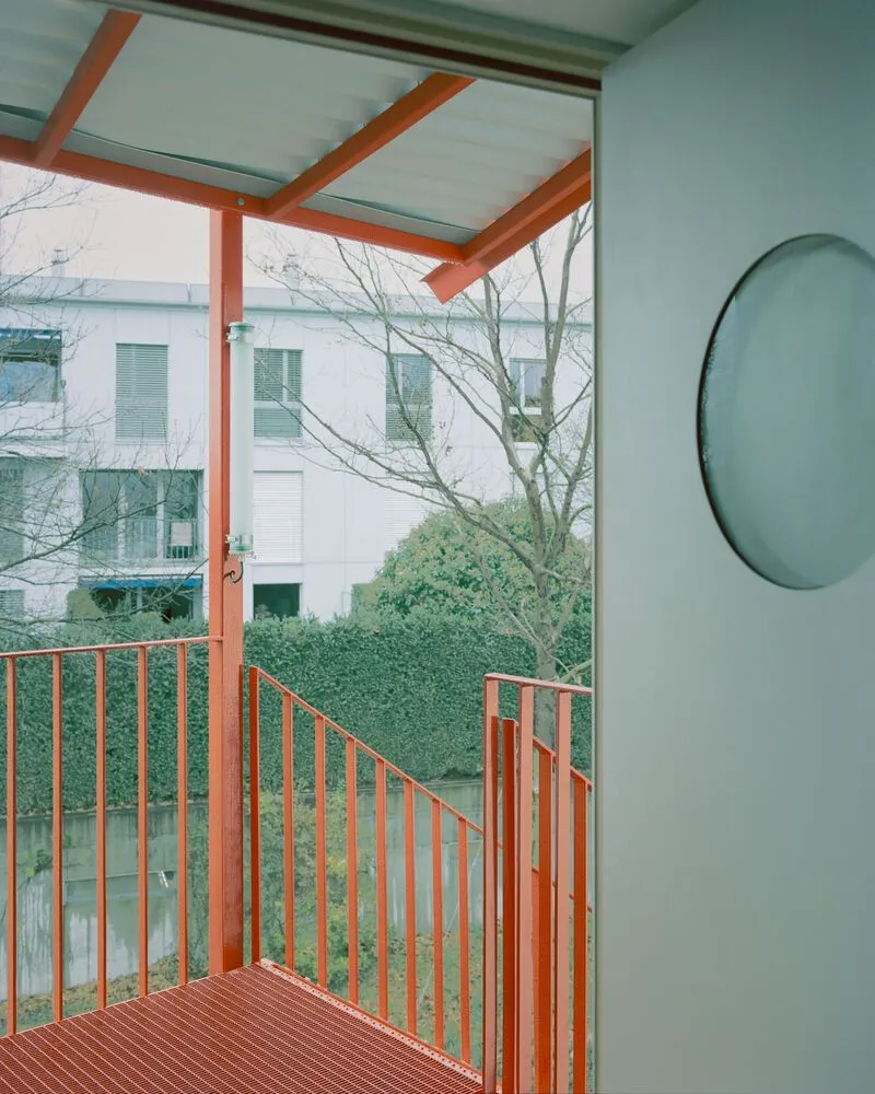 Orange steel balcony with vertical slat railing overlooking bare winter trees and neighboring buildings