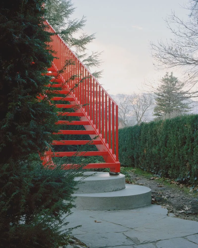 Orange steel exterior staircase emerging from evergreen trees along a paved garden path in winter