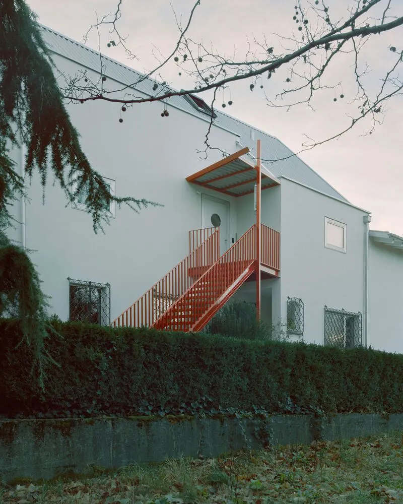 White rendered facade with red metal exterior staircase and balcony beneath bare winter branches