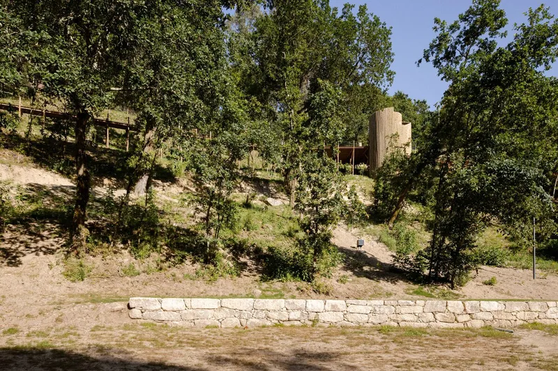 Distant view of the cylindrical timber tower sitting on a wooded hillside above a stone retaining wall