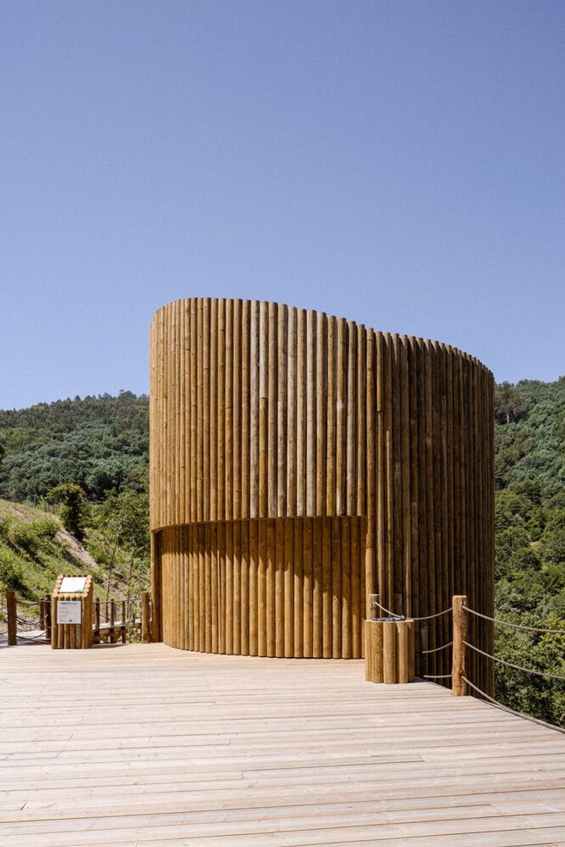 Curved timber-slatted tower rising from a wooden deck with forested hills beyond in afternoon light