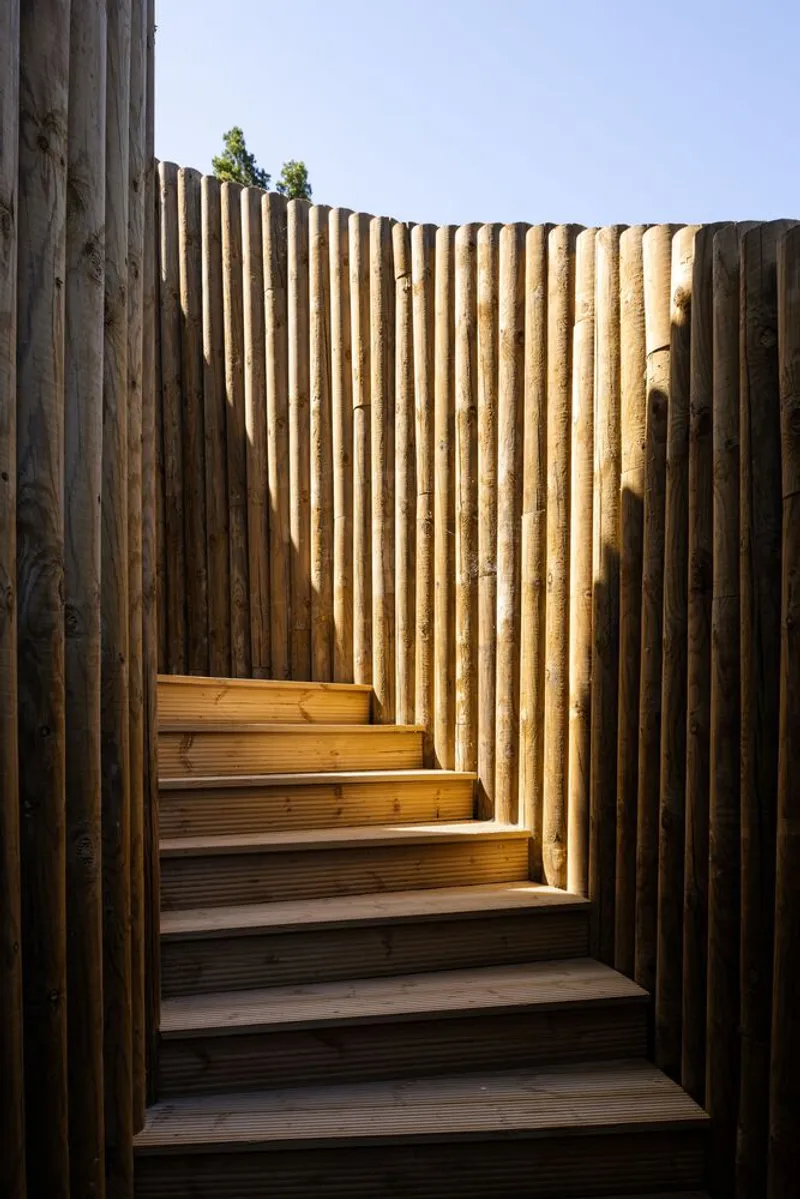 Open-air timber staircase ascending between weathered vertical wood column walls under clear sky