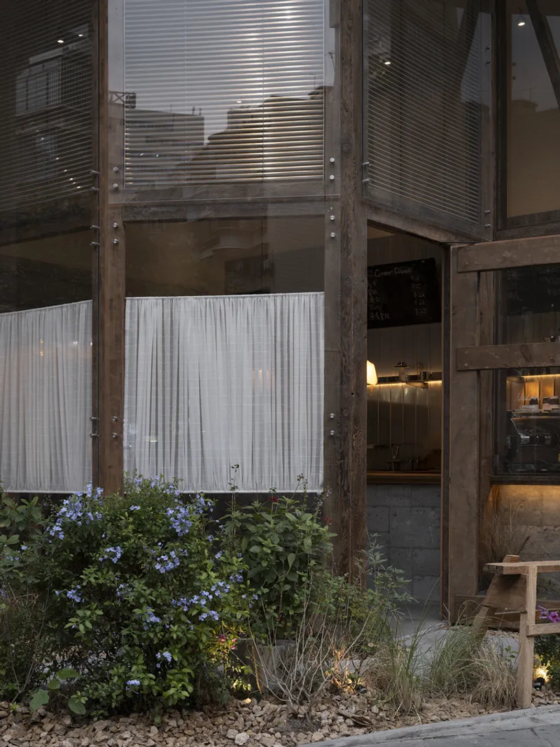 Street facade with timber frame, horizontal blinds, and planted bed with flowering shrubs at twilight