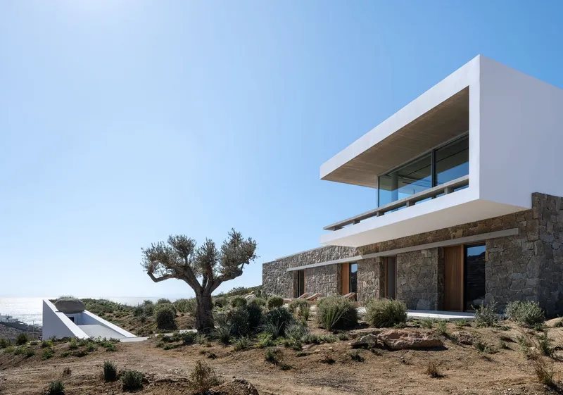 Stone-clad lower volume with timber openings beneath the white cantilevered upper floor and an olive tree