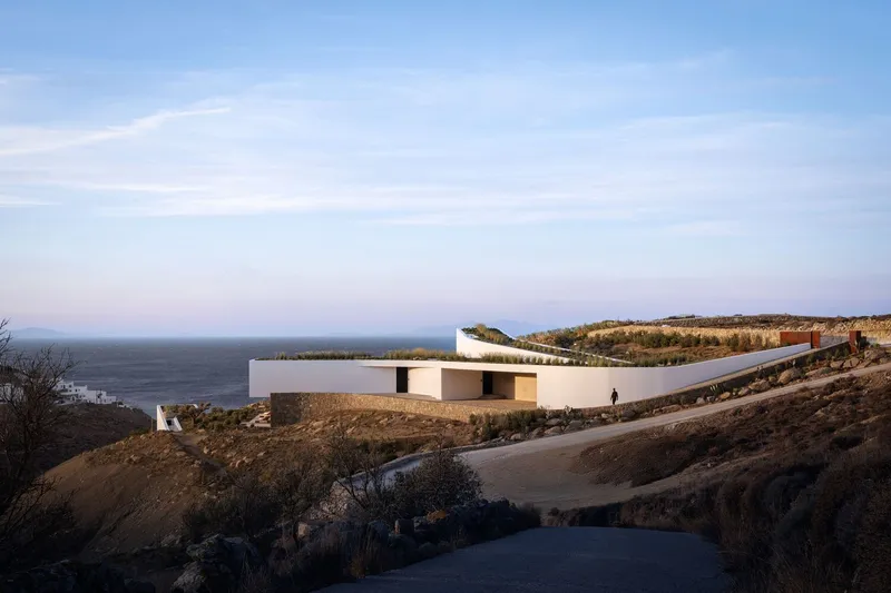 Distant view of the white volumes and cantilevered roof nestled into the rocky coastal hillside at dusk
