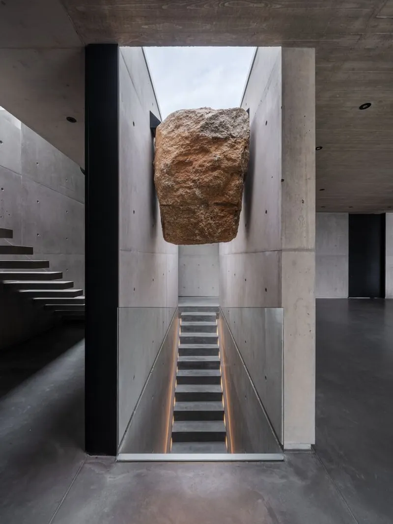 Large boulder suspended above a concrete stairwell with board-formed walls and natural light from above