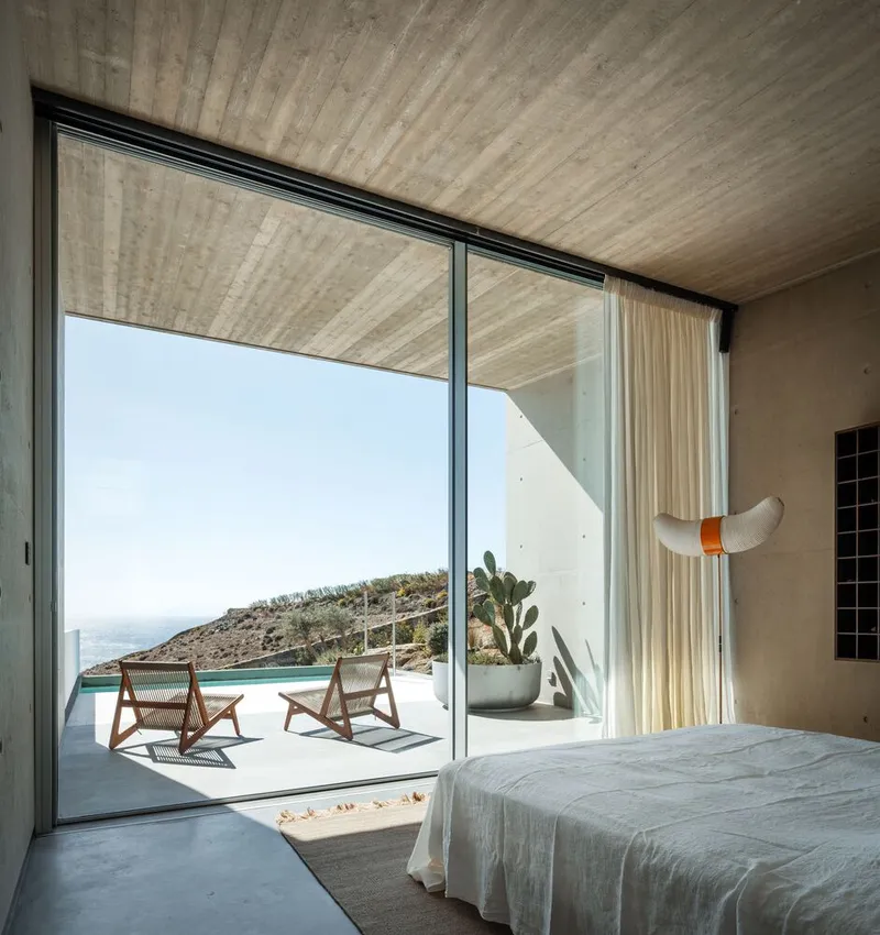 Bedroom opening to poolside terrace with lounge chairs and hillside cacti under clear sky