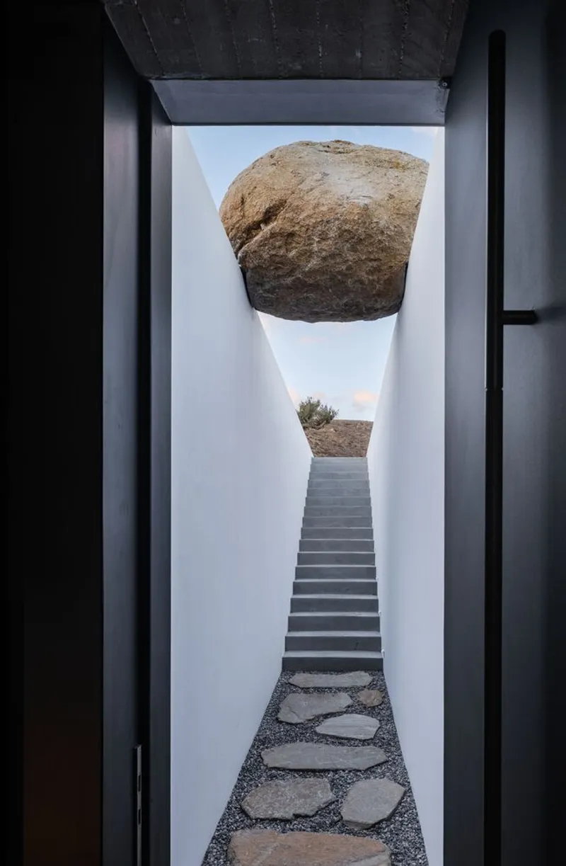 Courtyard corridor with stone paver path leading to stairs beneath suspended boulder and sky