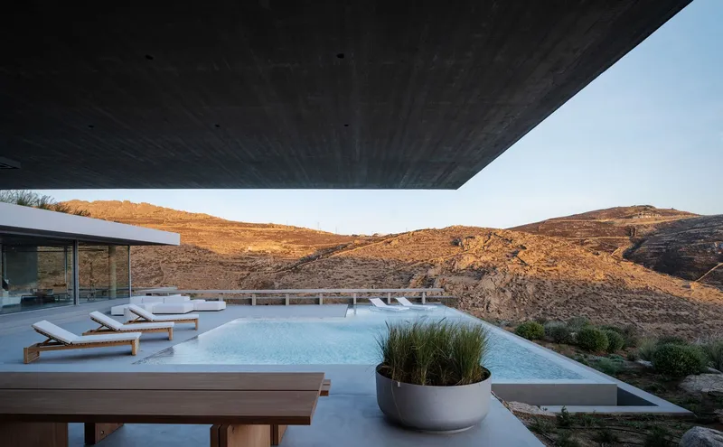 Infinity pool terrace with timber loungers beneath a cantilevered soffit facing rocky hills at sunset