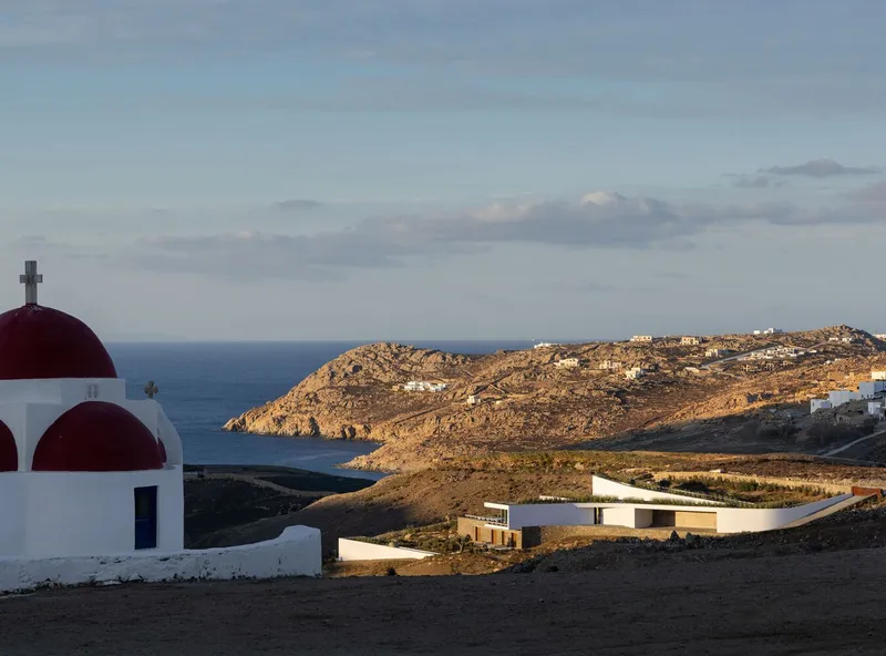 White chapel with red domes beside horizontal volumes nestled in a coastal landscape
