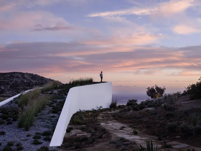 White cantilevered volume emerging from a rocky hillside with a figure on its roof at dusk