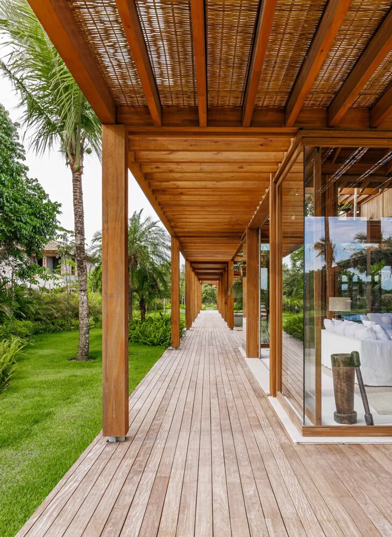 Covered timber walkway with exposed beams and woven ceiling panels alongside glass facades and palm trees