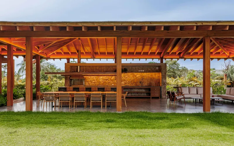 Open timber pavilion with exposed rafters sheltering an outdoor kitchen and dining area overlooking a lawn