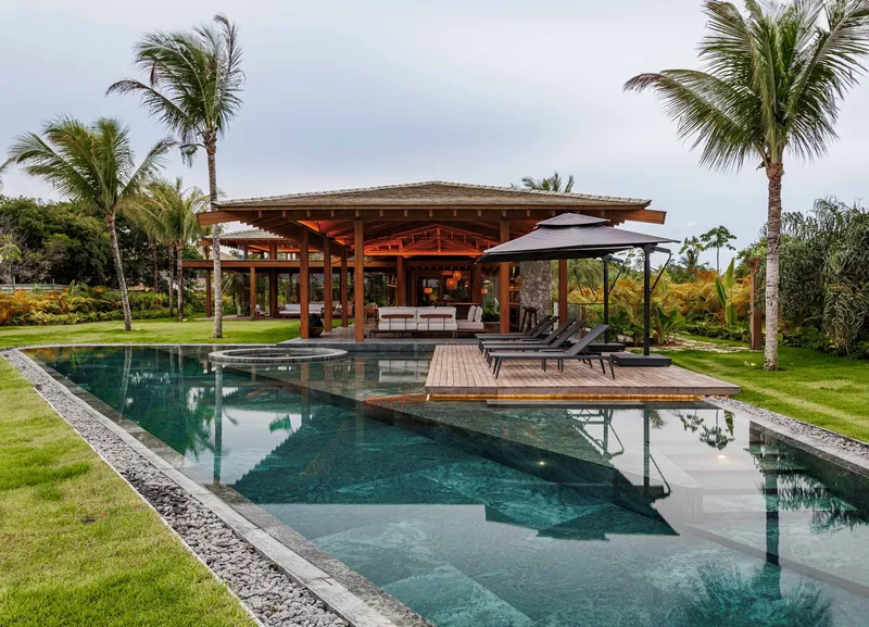 Pool terrace with timber deck islands and pavilion framed by palm trees under cloudy sky