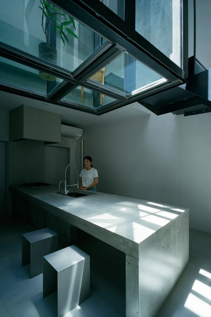 Concrete kitchen island beneath black steel skylight bridge with person working at the counter