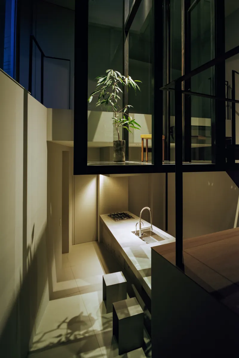 Night view through steel-framed glazing showing potted plant and kitchen counter with warm interior lighting