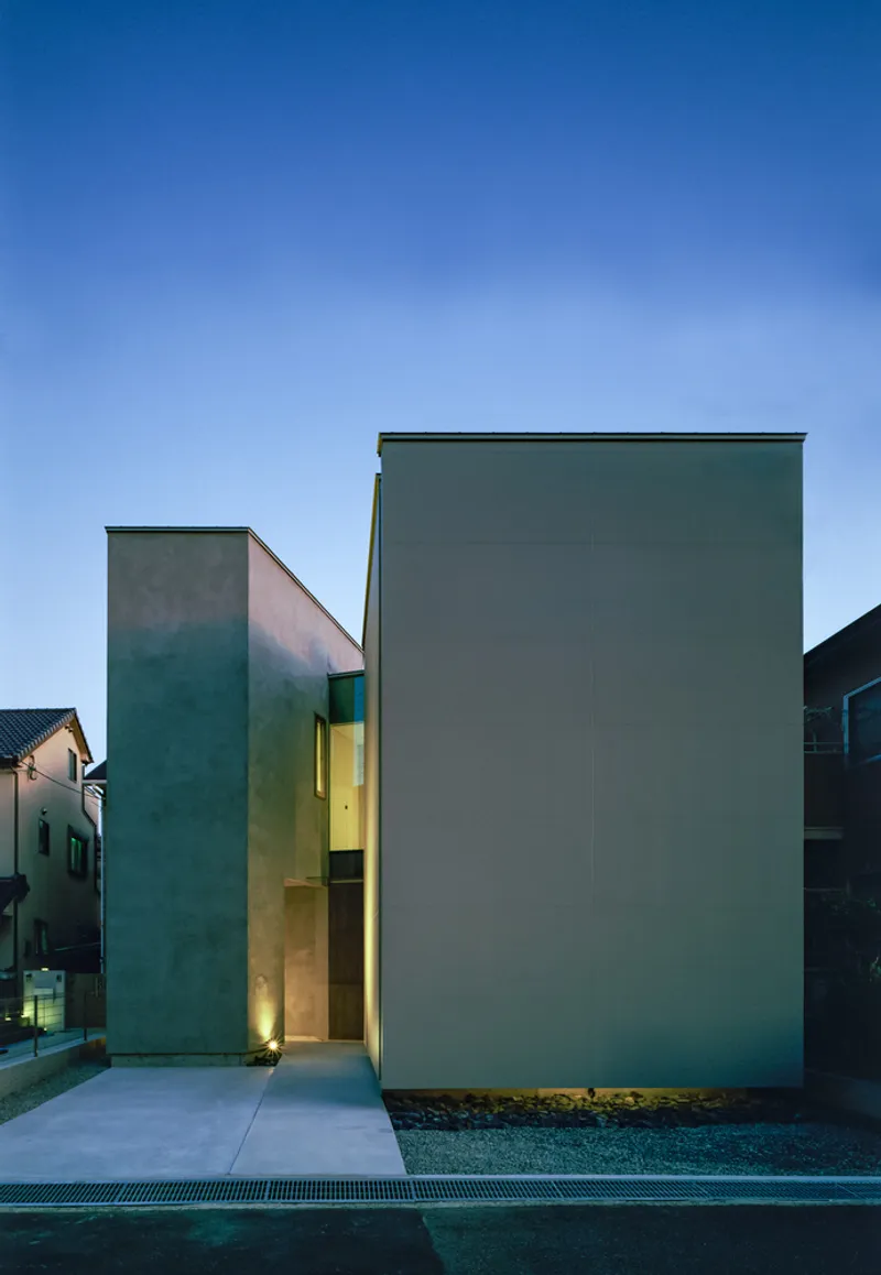 Street facade with two grey concrete volumes flanking a lit yellow entrance at dusk