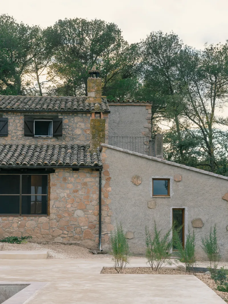 Exterior view of renovated stone house with clay tile roof and young planted grasses at dusk