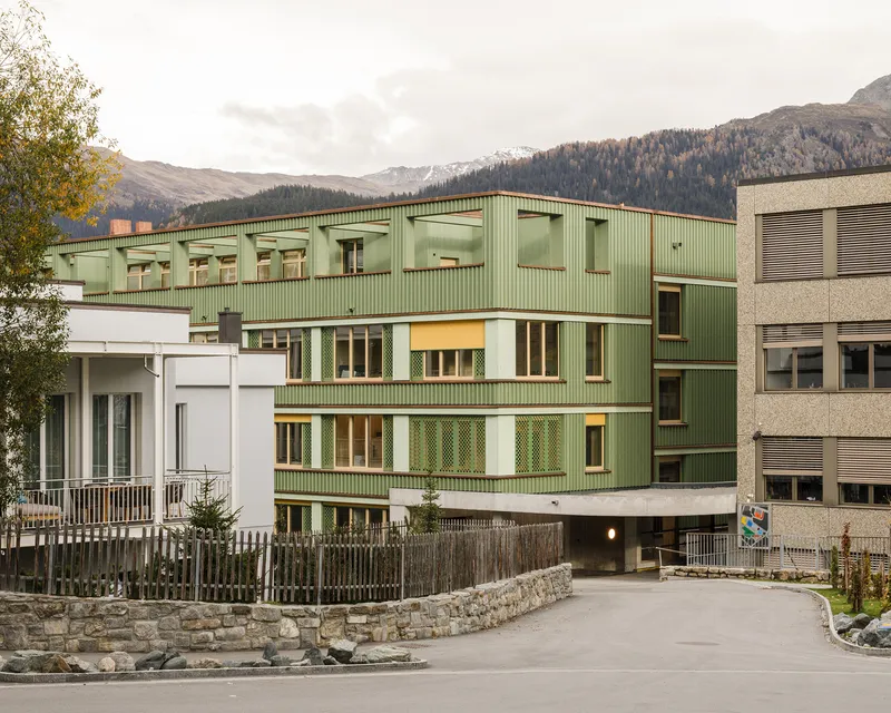 Multi-story green corrugated metal facade with horizontal band windows against a forested mountain backdrop