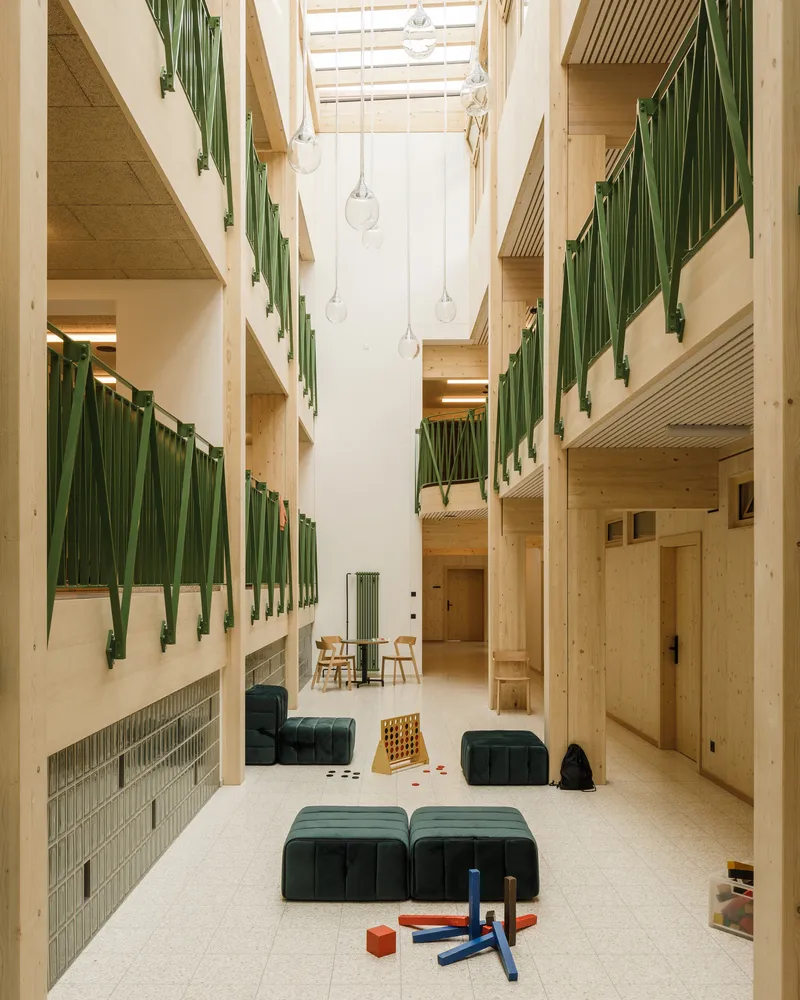 Triple-height atrium with exposed timber balconies, green metal railings, and pendant lights beneath a skylight