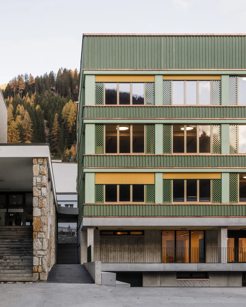Close-up of the green corrugated metal facade with perforated metal shutters flanking illuminated windows at dusk