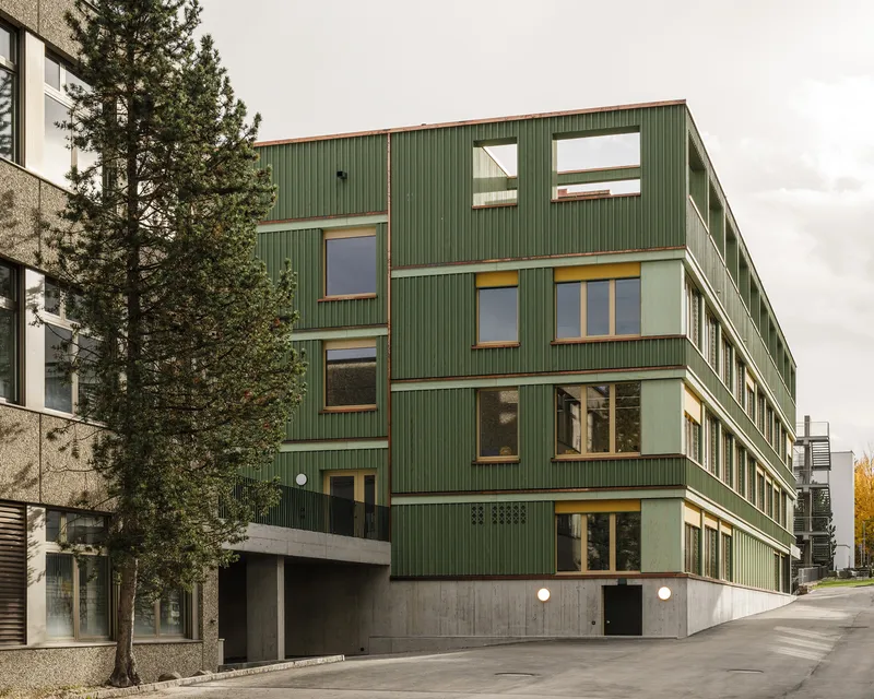 Street-facing corner of the green corrugated metal building with staggered window openings and concrete base
