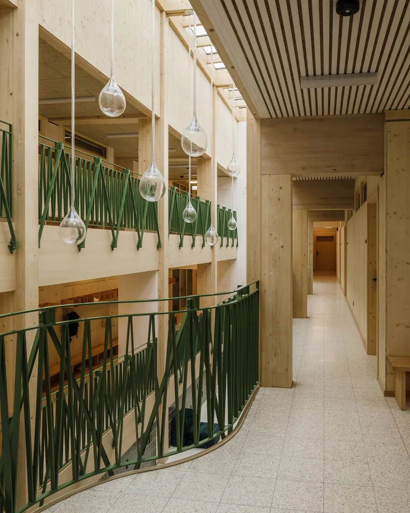 Double-height interior corridor with green metal balustrade and glass pendant lights suspended from timber ceiling