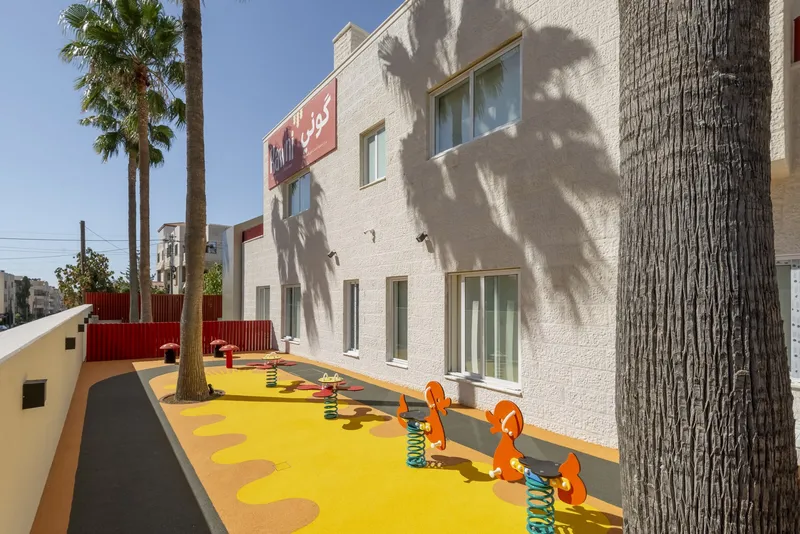 Outdoor play area with yellow rubber surfing, orange spring riders, and palm trees casting shadows on white stucco walls