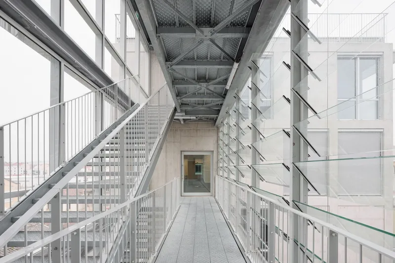 Elevated steel and glass walkway with perforated metal ceiling and railings spanning between building volumes