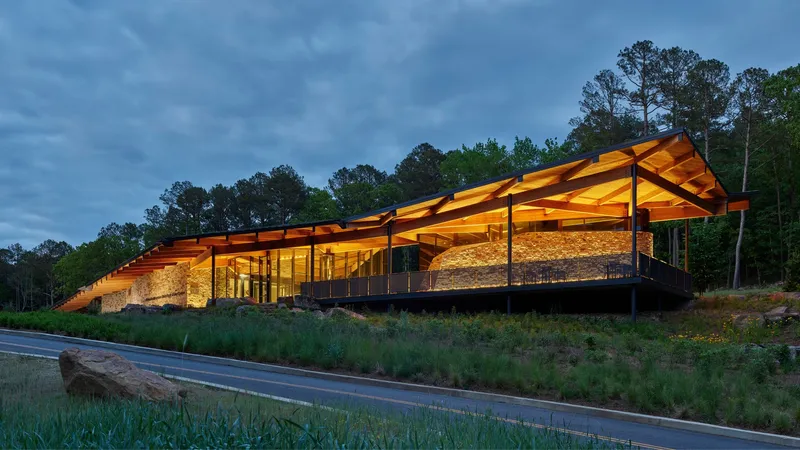 Illuminated visitor center with folded timber roof and stone walls at twilight beside a wooded hillside