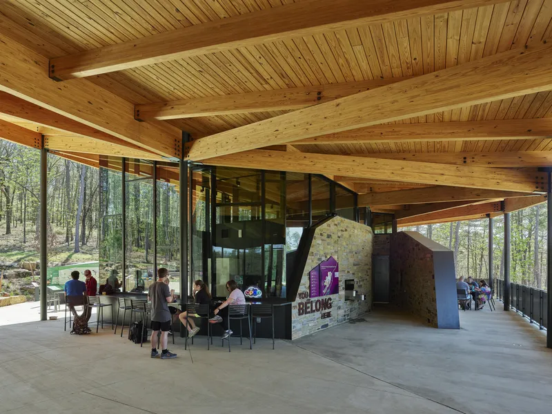 Open gathering area under curved timber beams with a fieldstone wall and visitors seated at tables