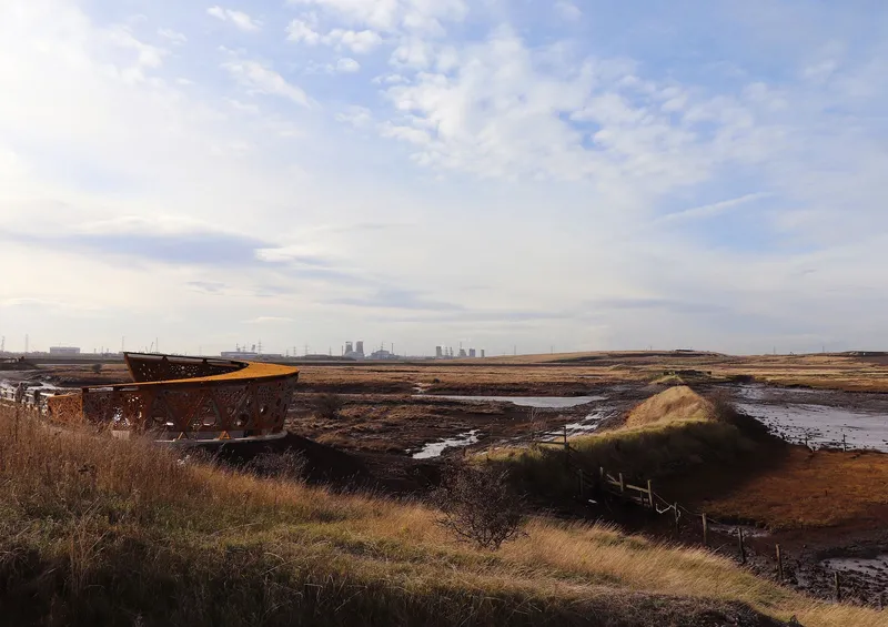Steel pavilion anchored in coastal grassland with industrial structures visible on the distant horizon