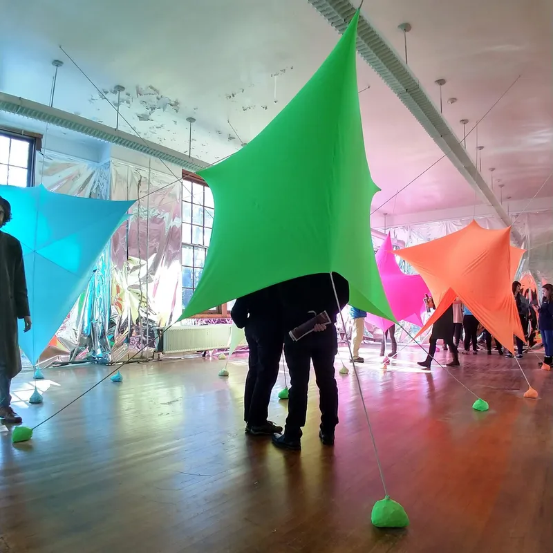 Gallery space with brightly colored fabric canopies anchored by green resin weights and visitors walking beneath