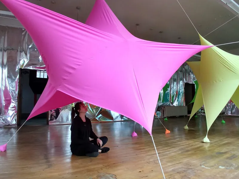 Person sitting beneath a pink tensile canopy with metallic curtain backdrop in gallery space