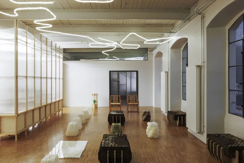 Open gallery space with corrugated furniture pieces scattered across oak floors and arched doorway in the background