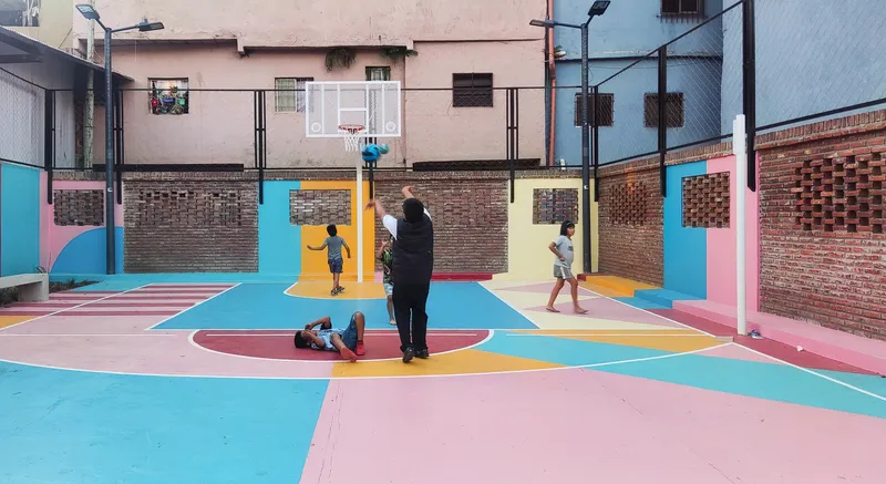 Outdoor basketball court with pink, blue and yellow painted surfaces surrounded by perforated brick walls
