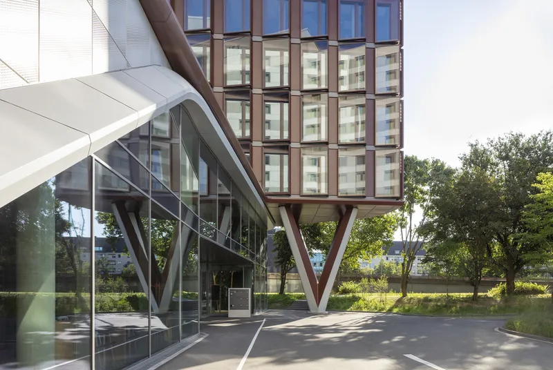 Entrance view showing the glazed ground floor and diagonal maroon structural columns supporting the elevated tower above