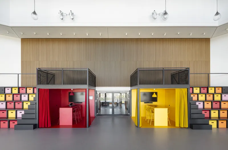 Interior atrium with two modular workstations in red and yellow beneath timber-clad mezzanines and colorful storage cubes